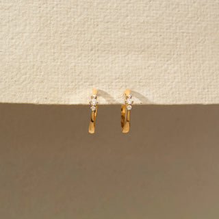 Front view of a pair of yellow gold hoop earrings accented with three graduated round diamonds on each hoop, suspended from a textured cream display surface.