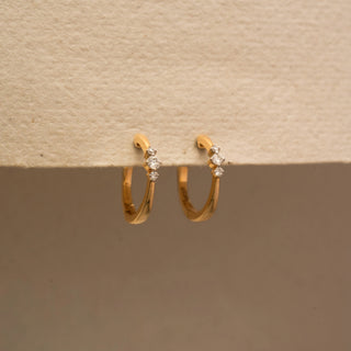 Side view of a pair of yellow gold hoop earrings featuring three graduated round diamonds on each hoop, suspended from a textured cream-colored display surface.