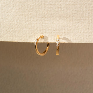 Angled view of a pair of yellow gold hoop earrings featuring three graduated round diamonds on each hoop, displayed on a textured cream-colored presentation surface.