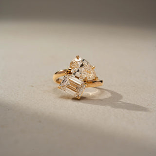 Front view of a toi et moi engagement ring in 14 KT Yellow Gold, featuring a pear-shaped and an emerald-cut lab-grown diamond in an elegant crossover setting, displayed against a beige backdrop.