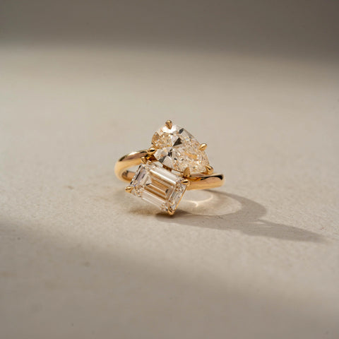 Front view of a toi et moi engagement ring in 14 KT Yellow Gold, featuring a pear-shaped and an emerald-cut lab-grown diamond in an elegant crossover setting, displayed against a beige backdrop.