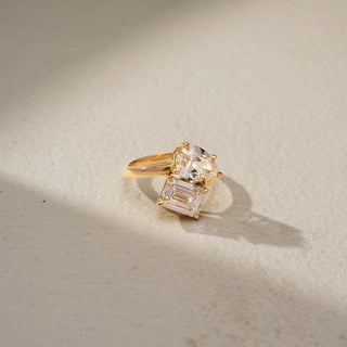 Top view of a toi et moi engagement ring in 14 KT Yellow Gold, featuring a pear-shaped and an emerald-cut lab-grown diamond in an elegant crossover setting, displayed against a beige backdrop.
