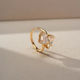 Angled view of a toi et moi engagement ring in 14 KT Yellow Gold, featuring a pear-shaped and an emerald-cut lab-grown diamond in an elegant crossover setting, displayed against a beige backdrop.