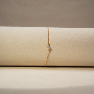 Front view of a solitaire diamond bracelet in 14 KT Yellow Gold, featuring a bezel-set round diamond on a delicate chain, displayed against a soft beige backdrop.