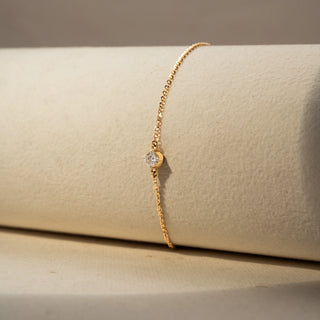 Side view of a solitaire diamond bracelet in 14 KT Yellow Gold, featuring a bezel-set round diamond on a delicate chain, displayed against a soft beige backdrop.