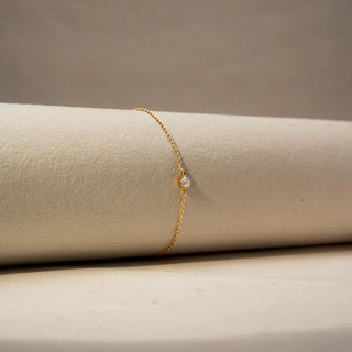 Angle view of a solitaire diamond bracelet in 14 KT Yellow Gold, featuring a bezel-set round diamond on a delicate chain, displayed against a soft beige backdrop.