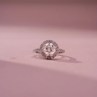 Front view of a round halo engagement ring in White Gold, featuring a round lab-grown diamond encircled by a sparkling halo and set on a pavé diamond band, displayed against a soft pink backdrop.
