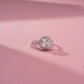 Side view of a round halo engagement ring in White Gold, featuring a round lab-grown diamond encircled by a sparkling halo and set on a pavé diamond band, displayed against a soft pink backdrop.