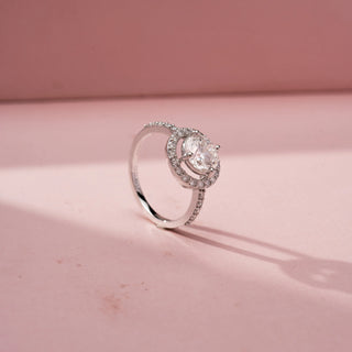 Angled view of a round halo engagement ring in White Gold, featuring a round lab-grown diamond encircled by a sparkling halo and set on a pavé diamond band, displayed against a soft pink backdrop.