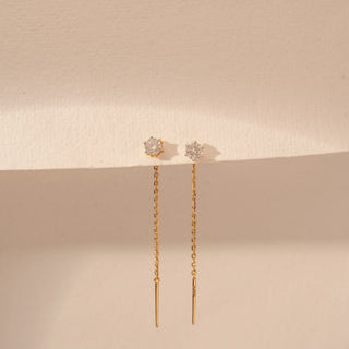 Front view of a 14KT yellow gold earrings featuring round-cut diamonds with delicate chain drops, set against a beige background.
