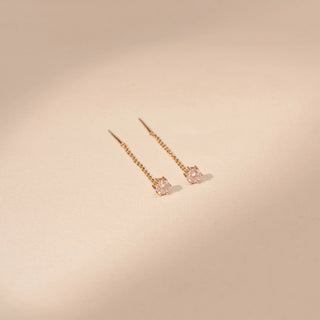 Side view of a 14KT yellow gold earrings featuring round-cut diamonds with delicate chain drops, set against a beige background.