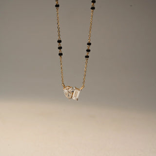 Front view of a two-stone diamond necklace in 14 KT Yellow Gold, featuring pear-shaped and emerald-cut lab-grown diamonds suspended from a delicate chain with black bead accents, displayed against a beige backdrop.