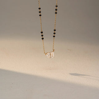 Side view of a two-stone diamond necklace in 14 KT Yellow Gold, featuring pear-shaped and emerald-cut lab-grown diamonds suspended from a delicate chain with black bead accents, displayed against a beige backdrop.