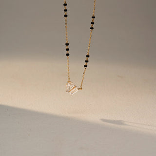 Angled  view of a two-stone diamond necklace in 14 KT Yellow Gold, featuring pear-shaped and emerald-cut lab-grown diamonds suspended from a delicate chain with black bead accents, displayed against a beige backdrop.
