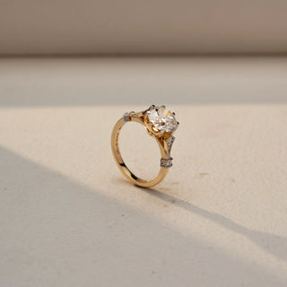 Angled view of a round solitaire engagement ring in 14 KT Yellow Gold, featuring a round lab-grown diamond accented by pavé-set diamond details along the cathedral-style shoulders, displayed against a beige backdrop.