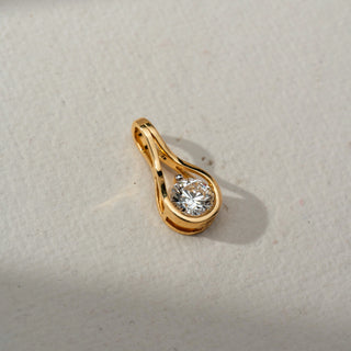 Top view of a gold pendant featuring a round brilliant-cut gemstone in a flowing teardrop-inspired setting, displayed on a soft neutral surface under natural light.