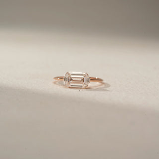 Front view of an emerald-cut solitaire ring in 14 KT Rose Gold, featuring an emerald-cut lab-grown diamond in a sleek east-west setting, displayed against a beige backdrop.