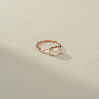 Side view of an emerald-cut solitaire ring in 14 KT Rose Gold, featuring an emerald-cut lab-grown diamond in a sleek east-west setting, displayed against a beige backdrop.