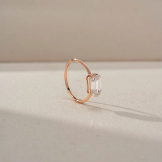 Angled view of an emerald-cut solitaire ring in 14 KT Rose Gold, featuring an emerald-cut lab-grown diamond in a sleek east-west setting, displayed against a beige backdrop.