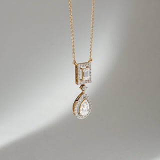 Angled view of Dual Drop Diamond Pendant Necklace in 14KT Yellow Gold, featuring an emerald-cut halo top with a pear-shaped lab grown diamond drop below. Set on a neutral backdrop, the design highlights depth and brilliance.
