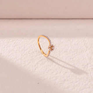 Side view of floral curve diamond ring in yellow gold, featuring a central flower cluster with round lab-grown diamonds and a slim pavé band. Set on a textured beige surface, the design casts a soft shadow.
