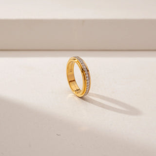 Angled view of a diamond band ring in 14KT Yellow Gold, featuring a row of round diamonds between polished gold borders against a beige backdrop.