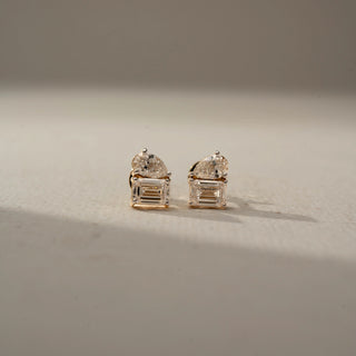 Front view of diamond stud earrings in yellow gold, featuring pear-shaped diamonds paired with emerald-cut diamonds. Displayed against a neutral backdrop, the contemporary design offers radiant sparkle.