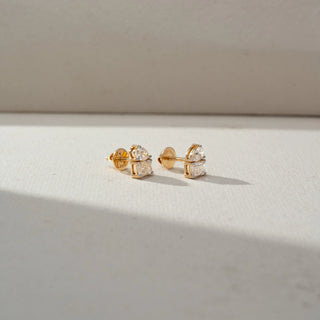 Side view of diamond stud earrings in yellow gold, featuring pear-shaped diamonds paired with emerald-cut diamonds. Displayed on a beige backdrop, the profile highlights the elegant dual-stone design.