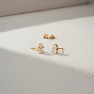 Angled view of diamond stud earrings in yellow gold, featuring pear-shaped diamonds paired with emerald-cut diamonds. Displayed on a beige backdrop, the dual-stone design showcases refined brilliance.