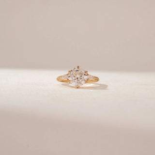 Front view of a round diamond ring in 14KT Yellow Gold, featuring a pavé band with intricate detailing against a beige backdrop.