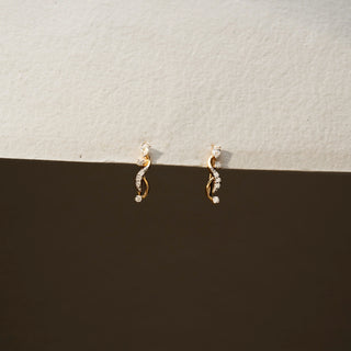 Front view of diamond swirl stud earrings in 14 KT Yellow Gold, featuring intertwined pavé-set curves accented with round diamonds against a neutral textured backdrop.