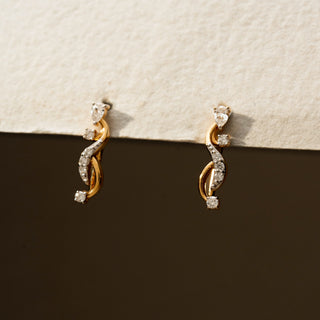 Close view of diamond swirl stud earrings in 14 KT Yellow Gold, featuring intertwined pavé-set curves accented with round diamonds against a neutral textured backdrop.