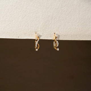 Side view of diamond swirl stud earrings in 14 KT Yellow Gold, featuring intertwined pavé-set curves accented with round diamonds against a neutral textured backdrop.
