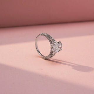 Angled view of an oval three-stone engagement ring in White Gold, featuring an oval-cut lab-grown diamond flanked by tapered baguette side diamonds and a pavé-set band, displayed against a soft pink backdrop