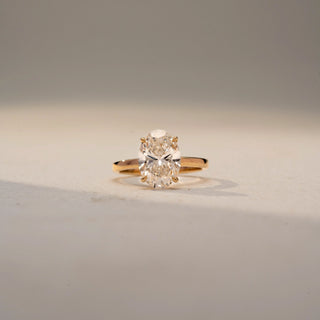 Front view of an oval solitaire ring in 14 KT Yellow Gold, featuring a prominent oval-cut lab-grown diamond in a classic four-prong setting, displayed against a beige backdrop.
