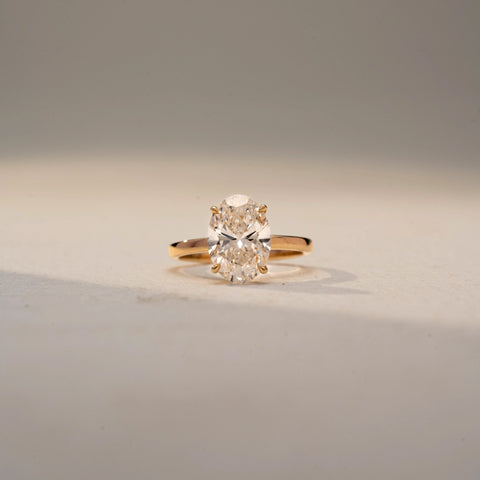 Front view of an oval solitaire ring in 14 KT Yellow Gold, featuring a prominent oval-cut lab-grown diamond in a classic four-prong setting, displayed against a beige backdrop.
