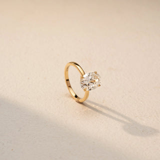 Angled view of an oval solitaire ring in 14 KT Yellow Gold, featuring a prominent oval-cut lab-grown diamond in a classic four-prong setting, displayed against a beige backdrop.