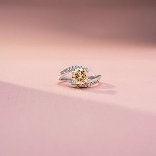 Front view of a bypass diamond ring in White Gold, featuring a round champagne-colored center diamond embraced by pavé-set diamond bands in a flowing crossover design, displayed against a soft pink backdrop.