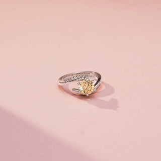 Side view of a bypass diamond ring in White Gold, featuring a round champagne-coloured centre diamond embraced by pavé-set diamond bands in a flowing crossover design, displayed against a soft pink backdrop.