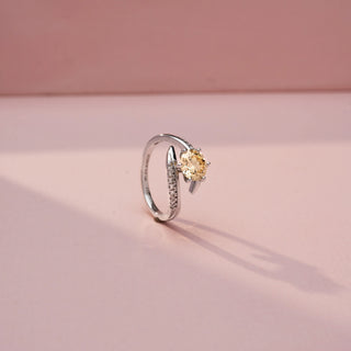 Angled view of a bypass diamond ring in White Gold, featuring a round champagne-coloured centre diamond embraced by pavé-set diamond bands in a flowing crossover design, displayed against a soft pink backdrop.