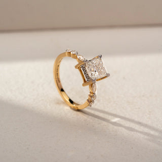 Angled view of a princess-cut solitaire engagement ring in 14 KT Yellow Gold, featuring a princess-cut lab-grown diamond accented by marquise-shaped side diamonds along the band, displayed against a beige backdrop.