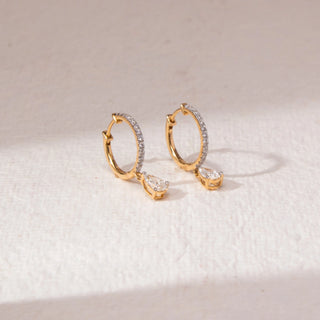 Angled view of diamond hoop drop earrings in yellow gold, featuring round lab-grown diamonds along the hoop with a pear-shaped diamond drop. Set on a soft neutral surface, they reflect delicate elegance and sparkle.