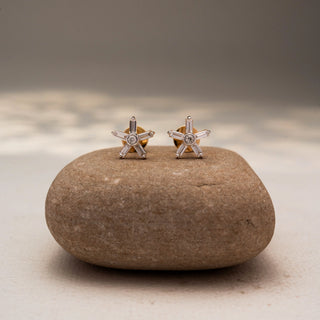 Front view of starburst diamond stud earrings in 14 KT Yellow Gold, featuring baguette and round diamonds arranged in a radiant six-point design, displayed on a textured stone surface.
