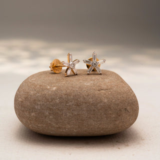 Side view of starburst diamond stud earrings in 14 KT Yellow Gold, featuring baguette and round diamonds arranged in a radiant six-point design, displayed on a textured stone surface.
