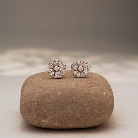 Front view of floral diamond stud earrings in 14 KT Yellow Gold, featuring pavé-set petal motifs radiating from a round center diamond, displayed on a textured stone surface.
