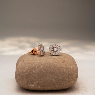 Angled view of floral diamond stud earrings in 14 KT Yellow Gold, featuring pavé-set petal motifs radiating from a round center diamond, displayed on a textured stone surface.
