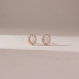 Front view of diamond stud earrings in 14KT yellow gold, featuring oval centre diamonds surrounded by pavé halo settings, displayed on a beige surface, highlighting brilliance and elegant symmetry.
