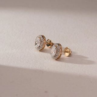 Side view of diamond stud earrings in 14KT yellow gold, featuring oval center diamonds with pavé halo settings and screw backs, displayed on a beige surface highlighting depth and brilliance.
