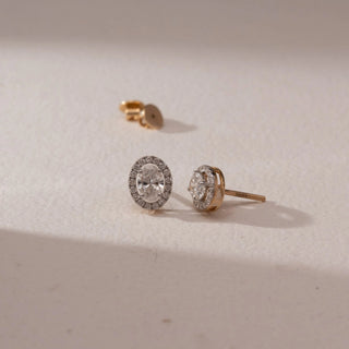 Angled view of diamond stud earrings in 14KT yellow gold, featuring oval center diamonds with pavé halo settings and screw backs, placed on a beige surface highlighting fine detail and sparkle.