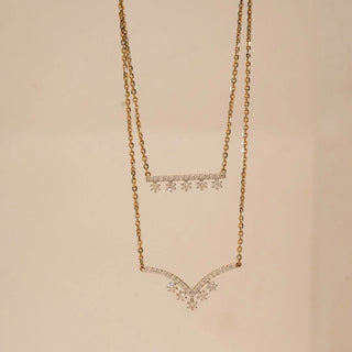 Front view of double layer yellow gold diamond necklace featuring a pavé diamond bar with dangling accents and a curved chevron diamond drop, displayed against a soft neutral background.